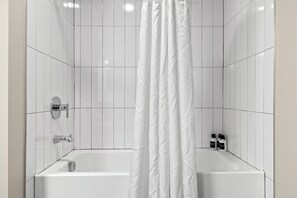 A bathroom with white tiled walls, a bathtub, a shower curtain, and two bottles of shampoo. - CozySuites at Westgate (Glendale)