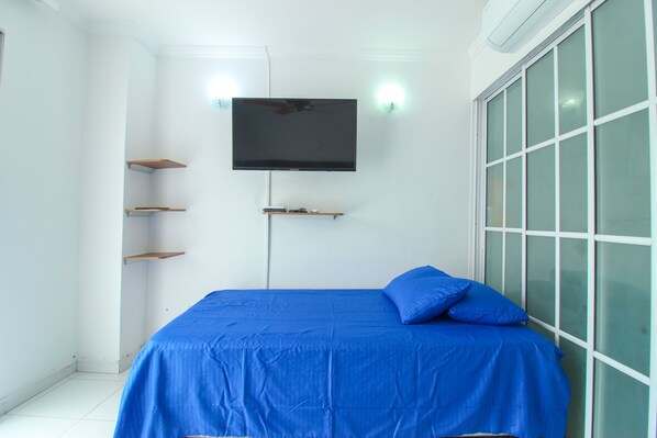 A bedroom with a bed, a TV mounted on the wall, and a window with blinds.