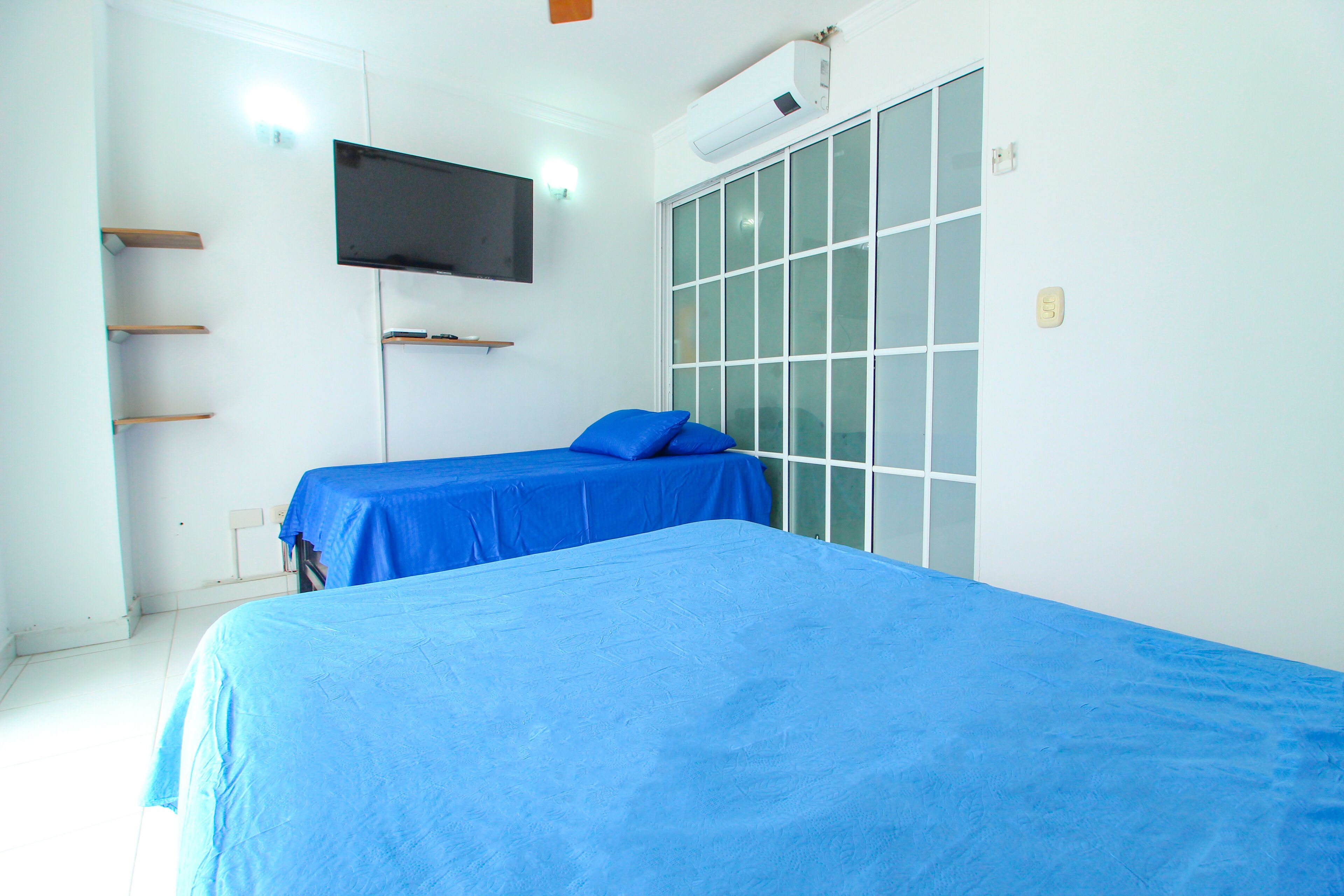 Quarto com duas camas, TV fixada na parede e porta de correr de vidro.