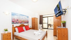 Une chambre bien rangée, avec un lit, une armoire en bois, un téléviseur fixé au mur et une grande fenêtre donnant sur l’extérieur.