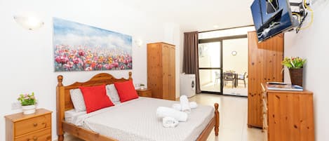 A neatly arranged bedroom with a bed, a wooden wardrobe, a television mounted on the wall, and a large window with a view of the outdoors.