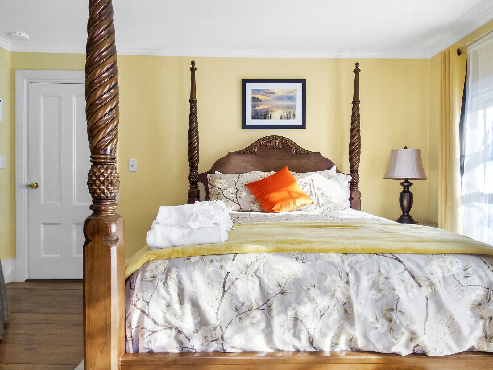 A four-poster bed with a floral bedspread, a framed picture above it, and a bedside lamp.