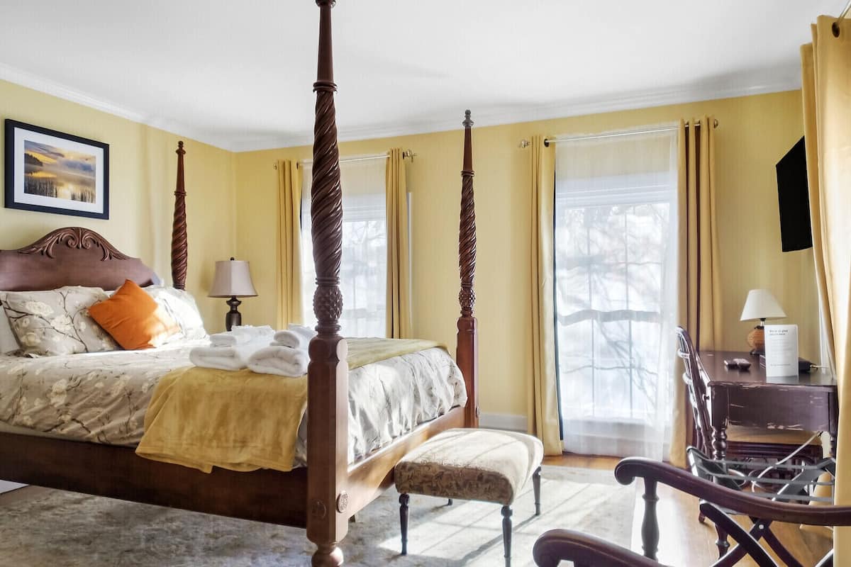 A four-poster bed with a headboard, a bedside table, a chair, and a desk with a lamp.