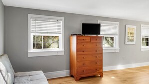 A room with a grey wall, wooden floor, a grey sofa, a wooden dresser, and white-framed windows with blinds. - The Thistle Inn (Boothbay Harbor)