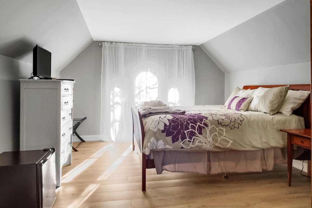 A bedroom with a bed, a dresser, a television on a cabinet, and a window with sheer curtains.