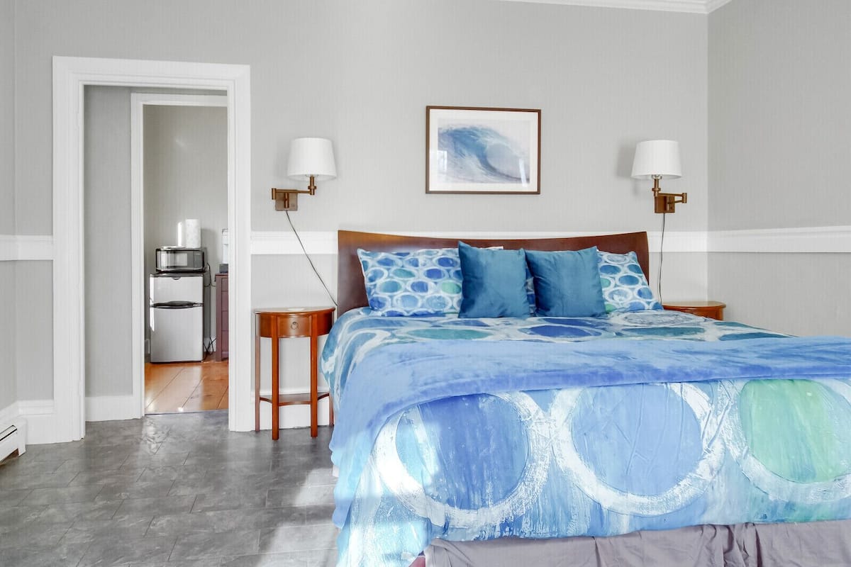A bedroom with a bed, two bedside tables, a nightstand, a wall-mounted lamp, and a framed picture.