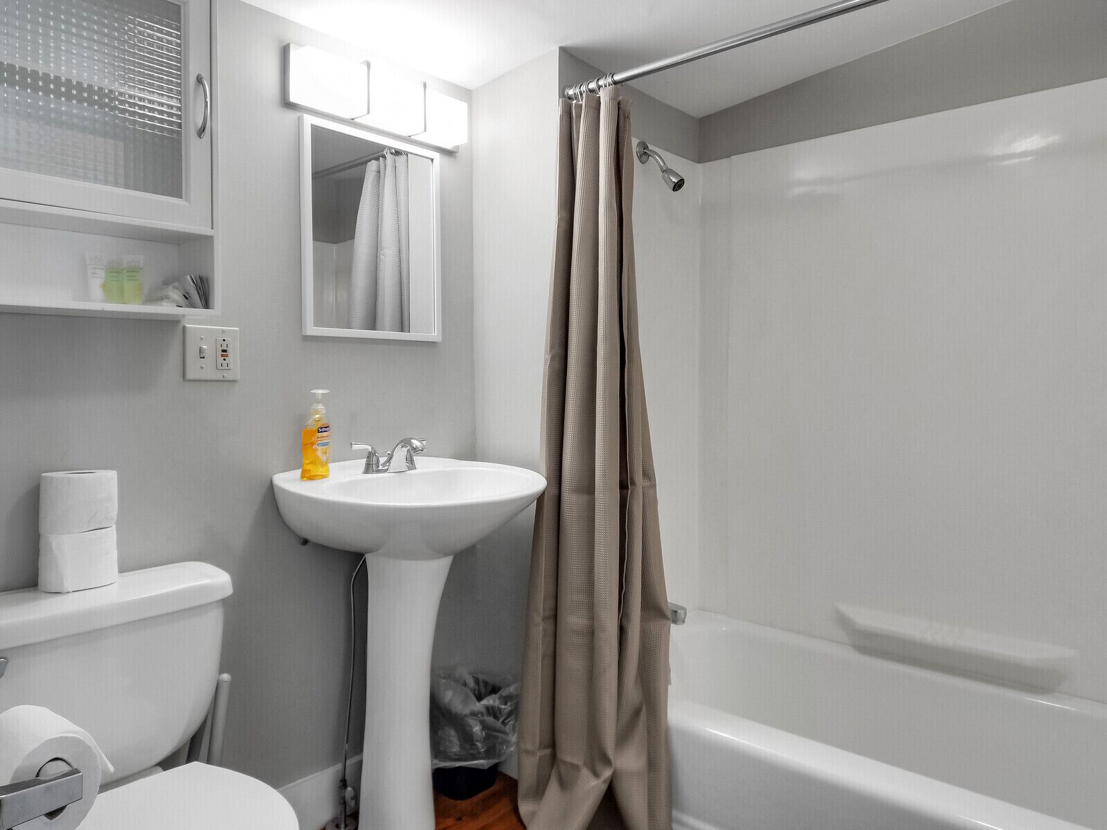 A bathroom with a white sink, toilet, and bathtub. There is a shower curtain, a mirror above the sink, and a shelf with toiletries.