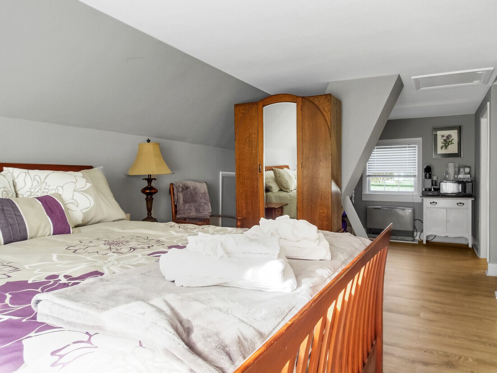 A bedroom with a bed, a nightstand, a lamp, a chair, a wardrobe, and a kitchenette.