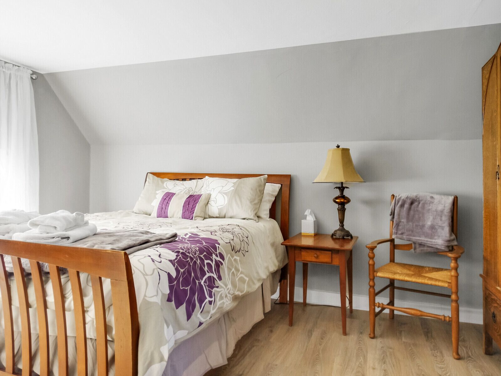 Ein ordentlich bezogenes Bett mit Holz-Kopfteil, ein Nachttisch mit Lampe und ein Sessel mit Decke.