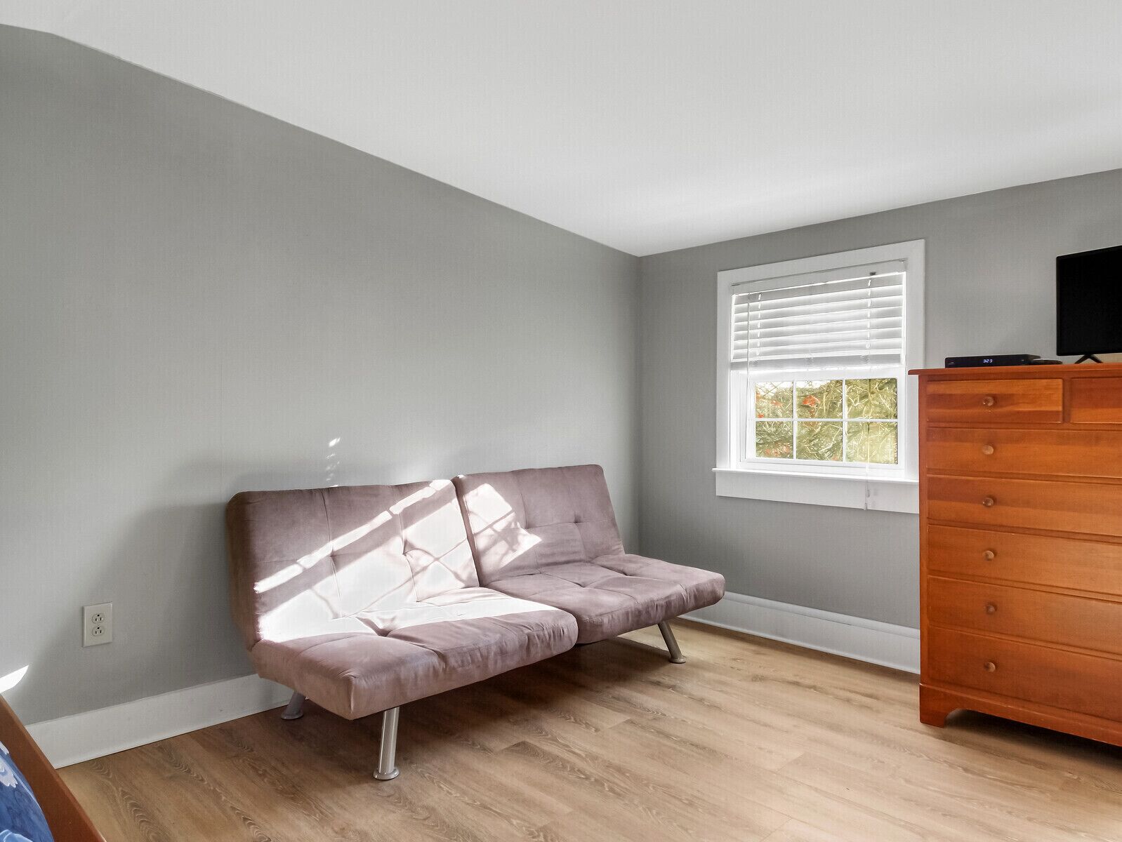 A room with a grey wall, a window with blinds, a wooden dresser, and a cushioned bench.