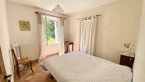 Ein Schlafzimmer mit einem Bett, einem Sessel, einer Heizung, einem Fenster mit Vorhängen, einem Deckenventilator und einem kleinen Tisch mit einer Lampe.