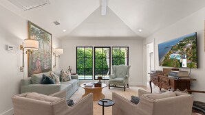 A spacious living room with a large window, a comfortable sofa, a coffee table, and a painting on the wall. - Quaint Cottage near the village with pool (St Simons Island)