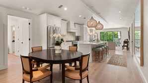 A spacious kitchen with white cabinetry, a central island with bar stools, and a dining area with a round table and chairs. - Quaint Cottage near the village with pool (St Simons Island)