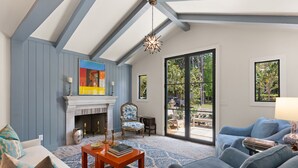 A living room with a fireplace, a blue sofa, a patterned rug, and a large window with a view of greenery. - Quaint Cottage near the village with pool (St Simons Island)