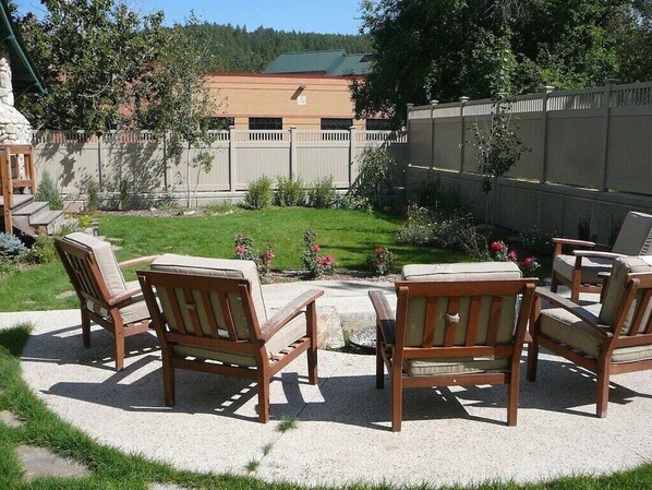 A patio area with four wooden chairs and a small table, surrounded by a concrete floor and a grassy lawn.