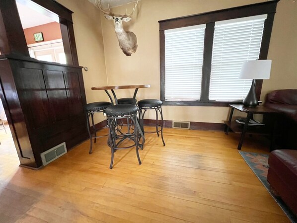 A room with a wooden floor, a table with four chairs, a mounted deer head on the wall, a window with blinds, and a lamp on a side table.