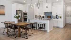 A modern kitchen with white cabinetry, stainless steel appliances, and a wooden dining table with wire chairs. - Lagoon Vibes: Desert Color Lagoon + Beaches (St George)