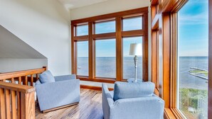 Quarto com uma grande janela com vista para o mar, com duas poltronas cinzentas e uma pequena mesa com abajur.