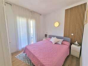Une chambre à coucher comprenant un lit, une table de chevet, une commode, une lampe fixée au mur et une fenêtre avec des rideaux.