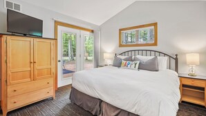 Een slaapkamer met een groot bed, een televisie, een kledingkast, een raam met uitzicht op bomen, en een nachtkastje met een lamp.