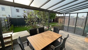 A covered patio area with a wooden table and chairs, a view of a backyard with a lawn and a tree, and a metal fence. - Modernes Reihenhaus mit Garten (Magdeburg)