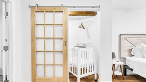 Ein Zimmer mit Holztür, einem weißen Babybett, einem kleinen Tisch mit Lampe und einem Bett mit weißer Bettwäsche.