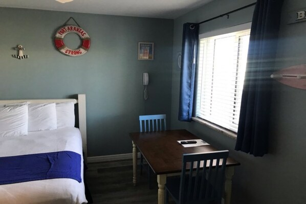 A room with a bed, a table, chairs, a window with curtains, a wall-mounted phone, and a life ring decoration. - Beachgate Condo Suites And Hotel 231+233 2 Bedroom Condo (Port Aransas)