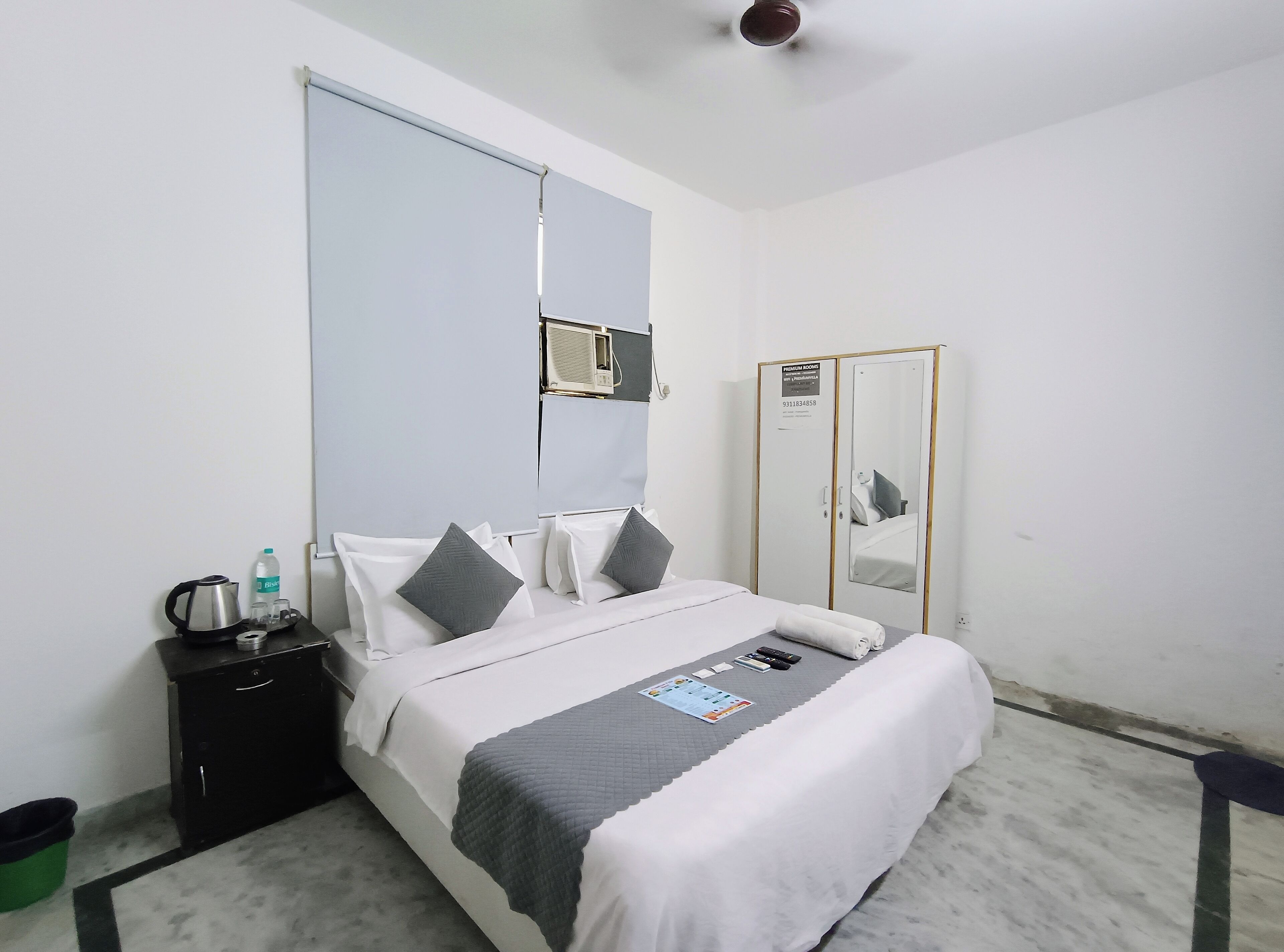 A hotel room with a bed, a bedside table with a kettle and a bottle, a mirror, and a wardrobe.