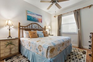 A bedroom with a bed, two bedside tables, a ceiling fan, a window with curtains, and a painting on the wall.