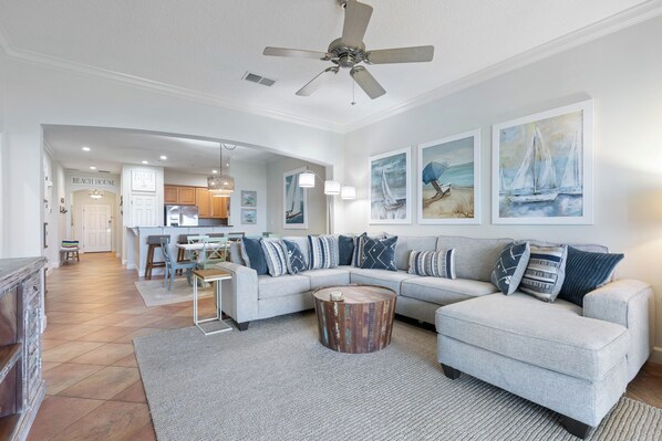 A spacious living room with a gray sectional sofa, a wooden coffee table, and a ceiling fan. A kitchen and dining area are visible in the background.