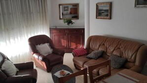 A living room with a brown leather sofa, two brown leather armchairs, a wooden coffee table, and a small side table. There are three framed pictures on the wall, a mirror above the cabinet, and a window with sheer curtains. - Piso As Rodas (Vilanova de Arousa)