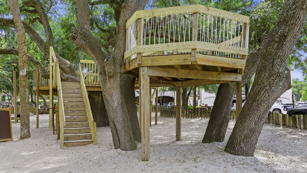 Una estructura de casa en árbol de madera con una escalera y una plataforma, rodeada de árboles y suelo arenoso.