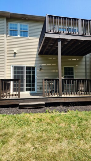 Une terrasse en bois avec des garde-corps, située devant un immeuble de deux étages doté de grandes fenêtres et d’une porte.