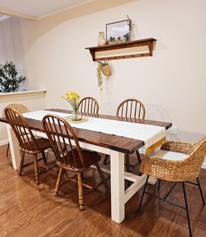 Une table à manger avec un plateau en bois et une nappe blanche, entourée de quatre chaises en bois et d’une chaise en osier. Une petite étagère au-dessus de la table expose des objets décoratifs.