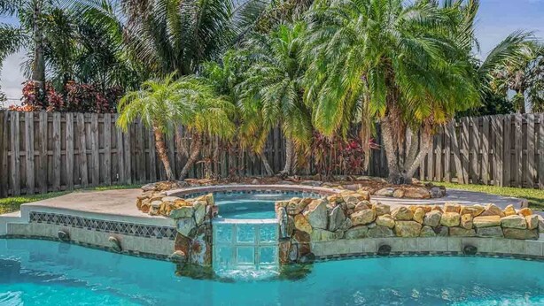 Piscina con cascata artificiale in pietra, circondata da palme e una recinzione in legno.