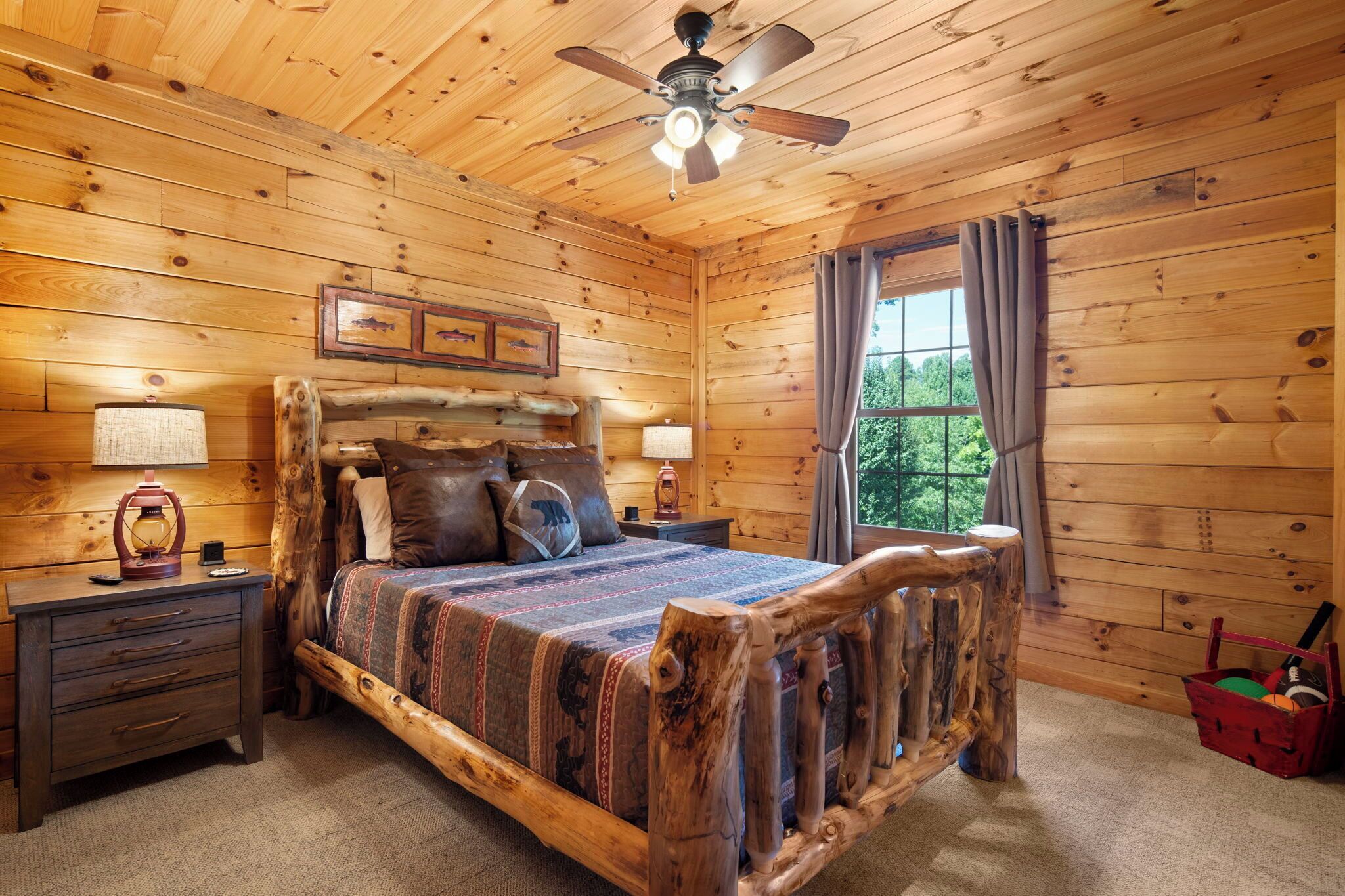A wooden cabin bedroom with a bed, nightstands, a ceiling fan, and a window with curtains.