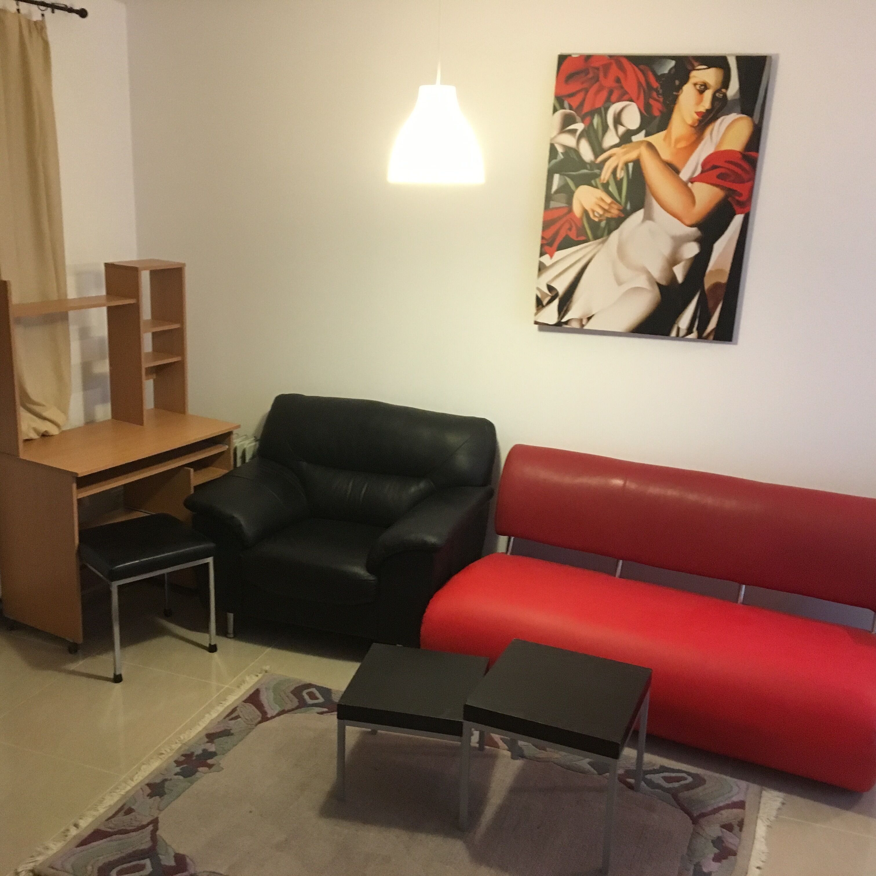 Un salon comprenant un fauteuil en cuir noir, un canapé rouge, une petite table noire, un bureau en bois avec des étagères et un tableau accroché au mur.