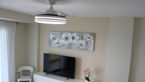 A modern living room with a flat-screen TV, a white cabinet, a ceiling fan, and a floral painting on the wall. - Don Ximo 13º  Solo Familias! (Playa de Gandía)
