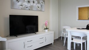 A modern living room with a flat-screen TV on a white cabinet, a dining table with white chairs, and a large floral painting on the wall. - Don Ximo 13º  Solo Familias! (Playa de Gandía)