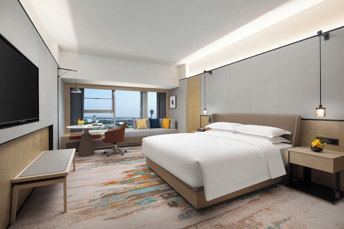 A modern hotel room with a large bed, a desk, a chair, and a view of the city.