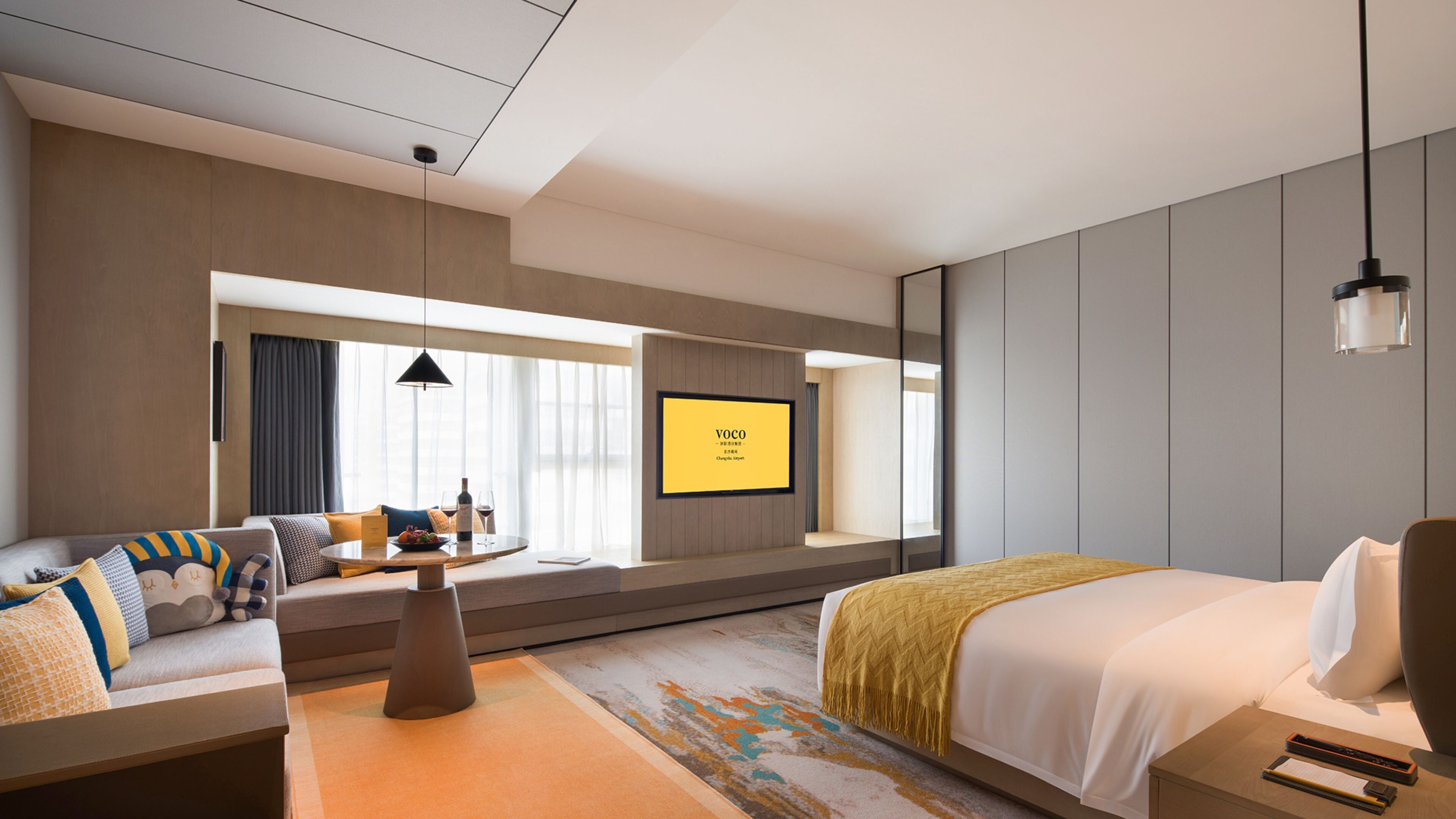 Una habitación de hotel moderna con una cama, una zona de estar con una mesa, un televisor montado en la pared y un ventanal grande.