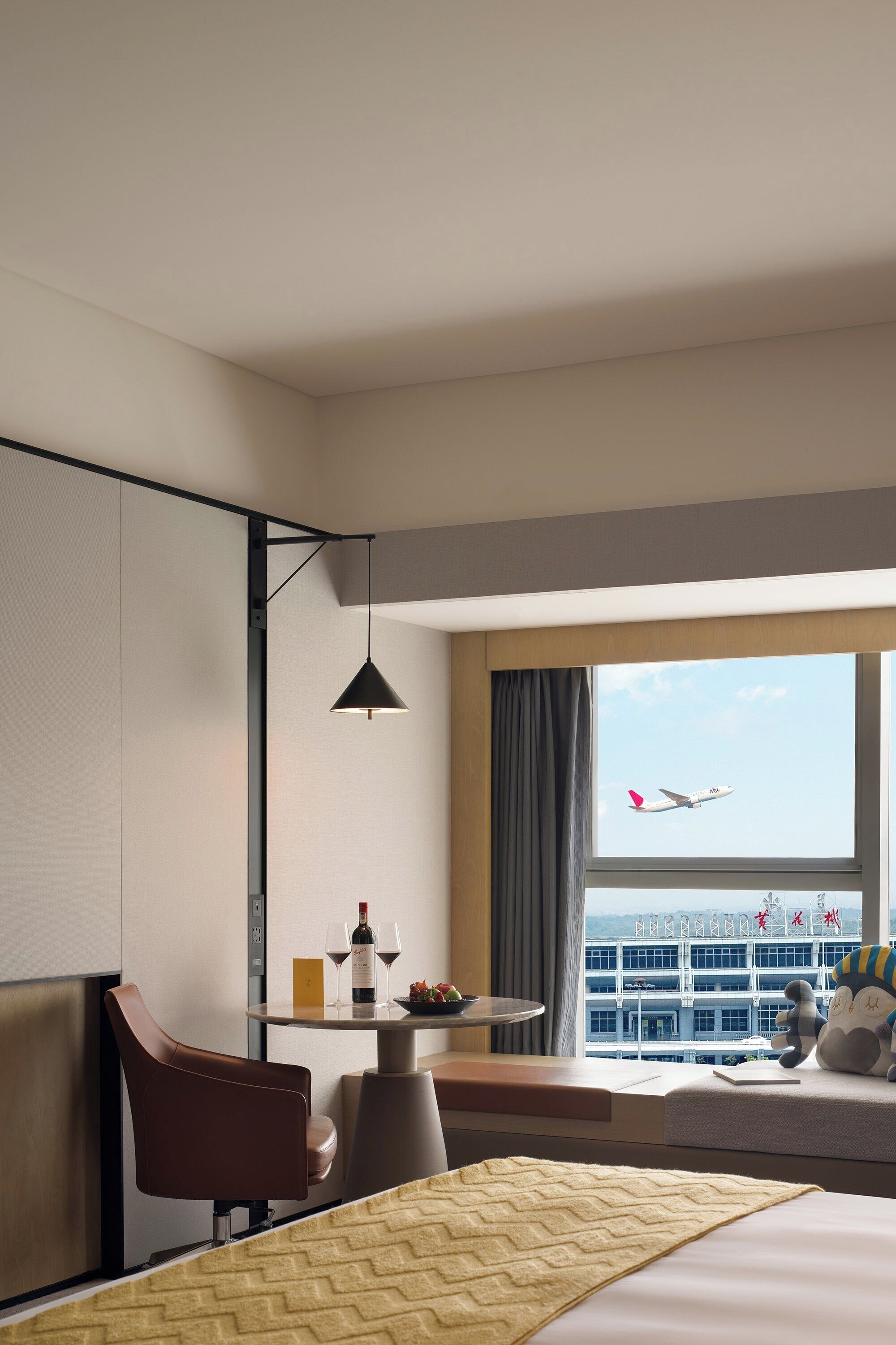Una camera d'albergo con un letto, una scrivania con una bottiglia e dei bicchieri, una sedia, una lampada a sospensione e una finestra con vista su un aereo.