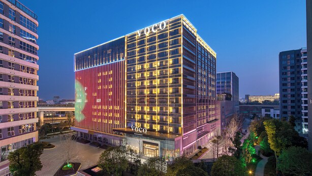 Ett modernt hotell med en tydlig skylt med texten ”Voco”, omgivet av andra byggnader och ett välskött grönområde.