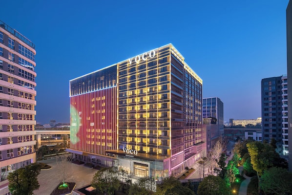 A modern hotel with a prominent 'Voco' sign, surrounded by other buildings and a landscaped area.