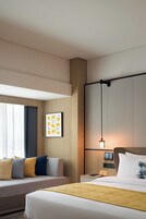 Habitación de hotel con cama, sofá, mesita de noche con lámpara, reloj digital y ventana con cortinas.