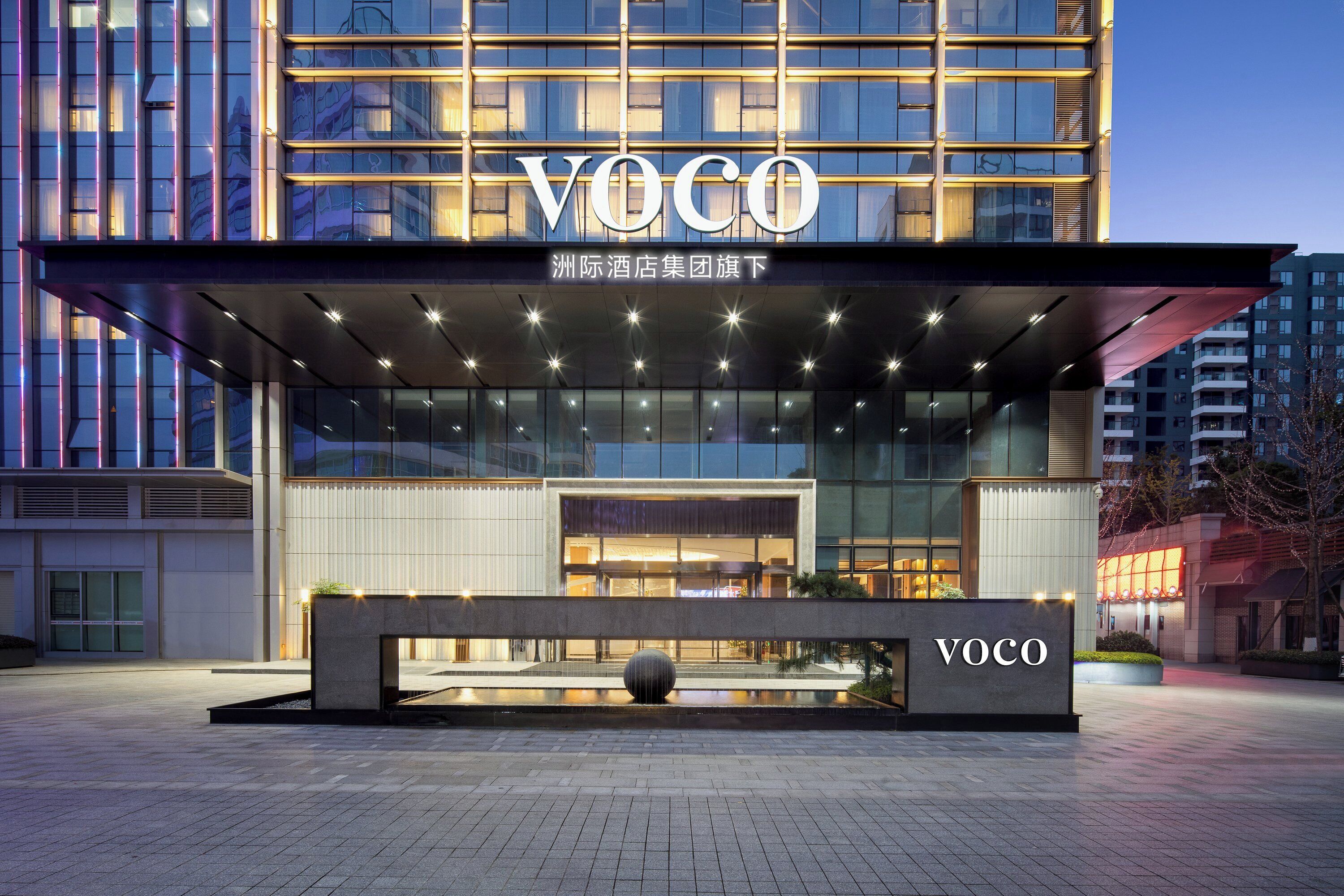 Ein moderner Hotelzugang mit einem auffälligen 'VOCO'-Schild, einem überdachten Außenbereich und einem Wasserspiel.