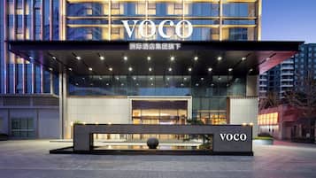 A modern hotel entrance with a prominent 'VOCO' sign, a covered outdoor area, and a water feature.