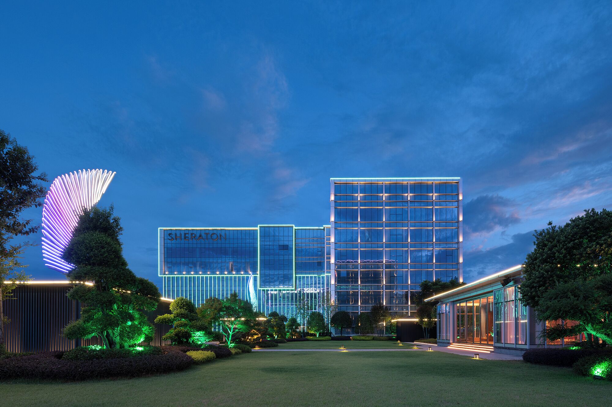 Un hotel moderno con fachada de vidrio, una estructura grande tipo ala y jardines bien iluminados.