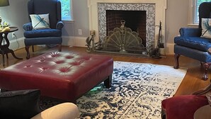 Un salon avec une cheminée, des fauteuils bleus, un pouf rouge et un tapis à motifs.