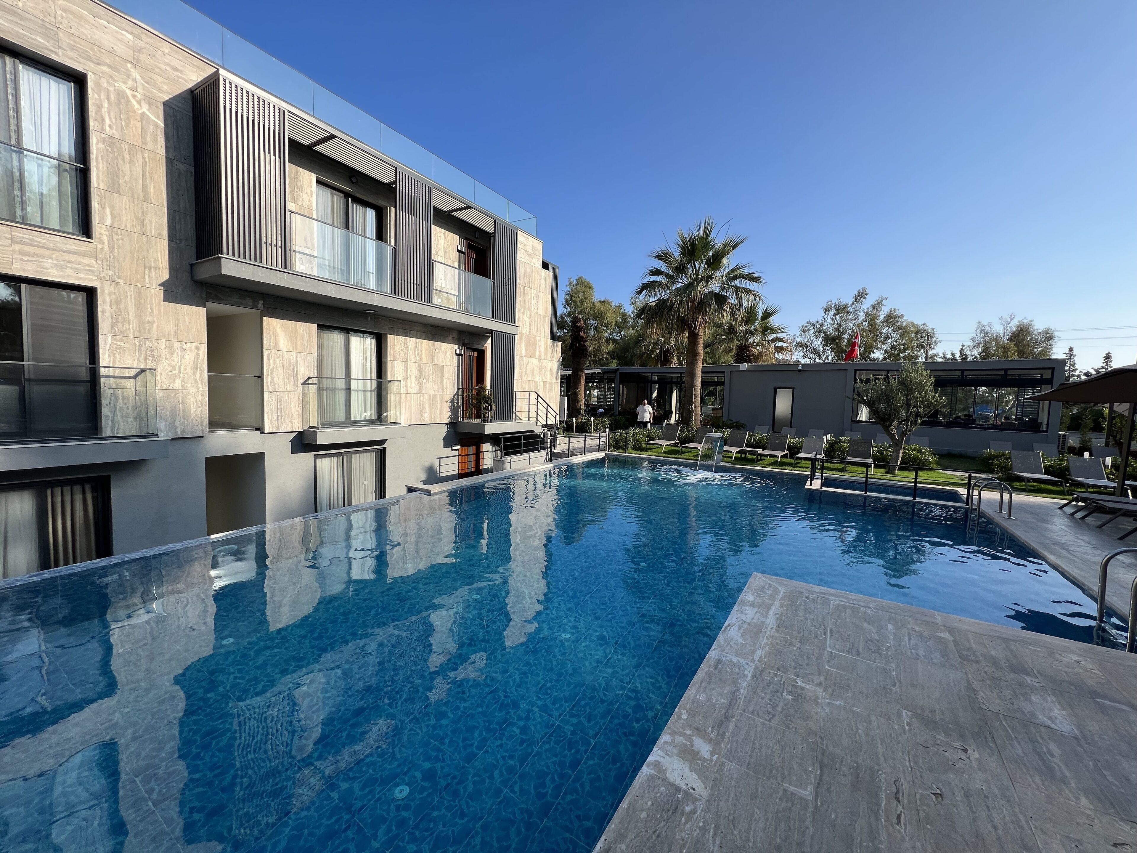 A modern hotel with a swimming pool, outdoor seating, and a clear blue sky.